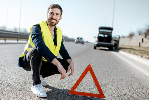 Portrait of a man near the emergency sign on the roadside with broken car on the background